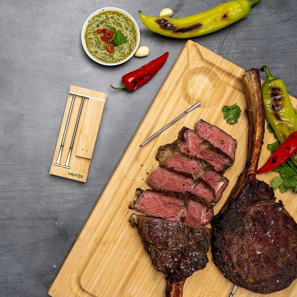 Sliced steak on a wooden cutting board with peppers and a small bowl of sauce on a gray surface using meater