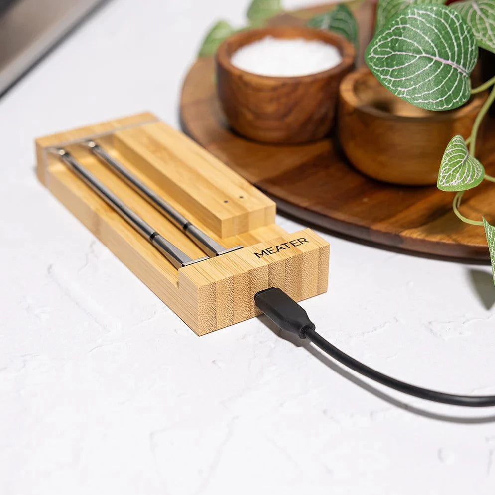 Wooden meat thermometer with 'MEATER' branding on a white surface with a wooden tray and plants in the background.