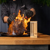 Person cooking a large piece of meat over flames with a MEATER device on a wooden surface.