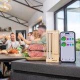 Meater digital meat thermometer on a kitchen counter with smartphone app displayed, people in the background.